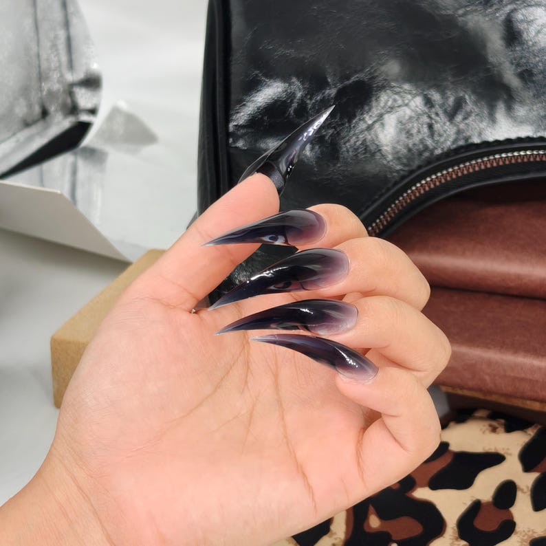 May include: A hand displaying long, pointed, black and purple ombre acrylic nails. The nails have a glossy finish and a sharp, stiletto shape. The nails are the focal point, with a blurred background.