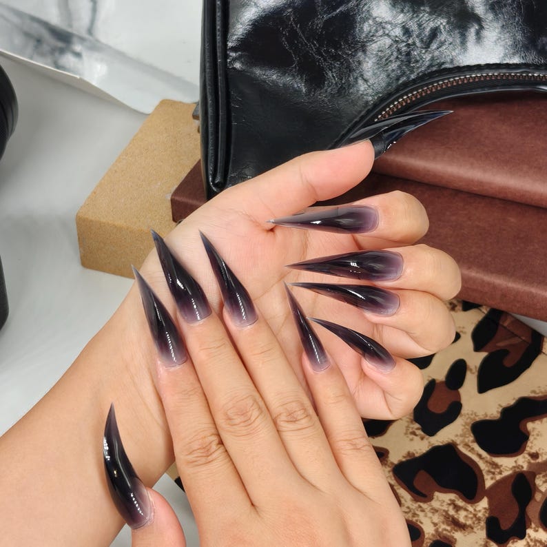 May include: A hand displaying long, stiletto-shaped artificial nails. The nails transition from a clear base to a dark, smoky gradient at the tips. The nails are glossy and reflect light, creating a dramatic effect. The overall aesthetic is bold and edgy.