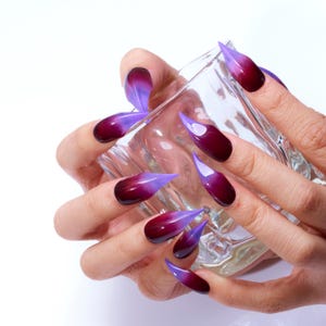 May include: Close-up of hands with long, pointed nails painted with a gradient of purple and dark red.