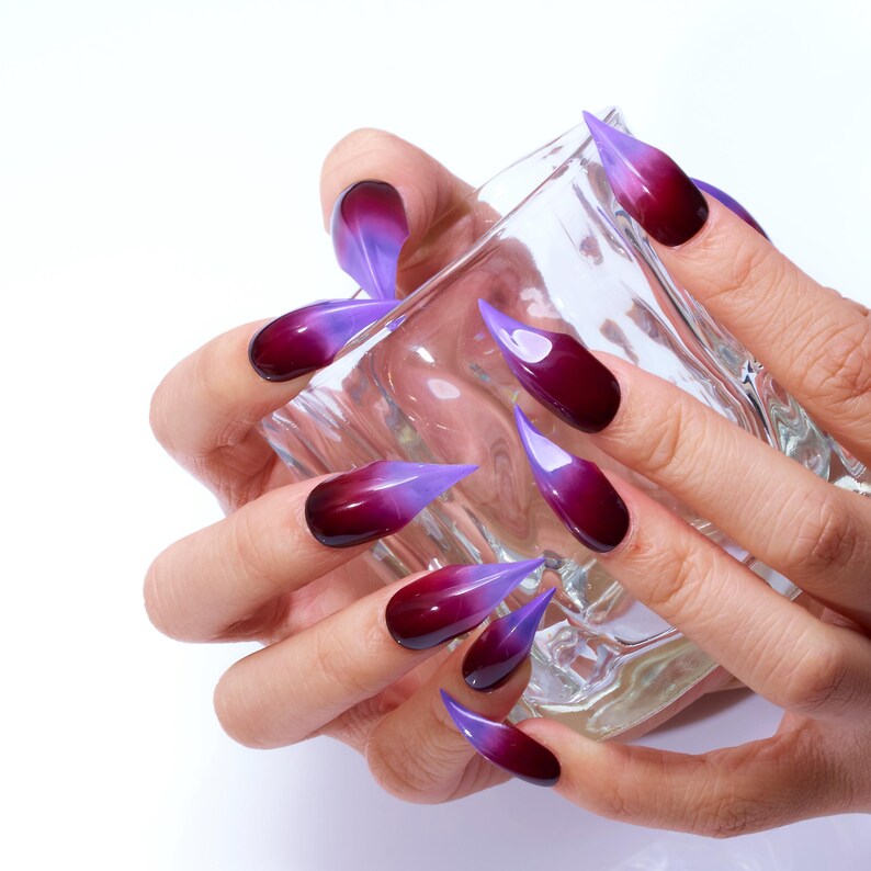 May include: Close-up of hands with long, pointed nails painted with a gradient of purple and dark red.