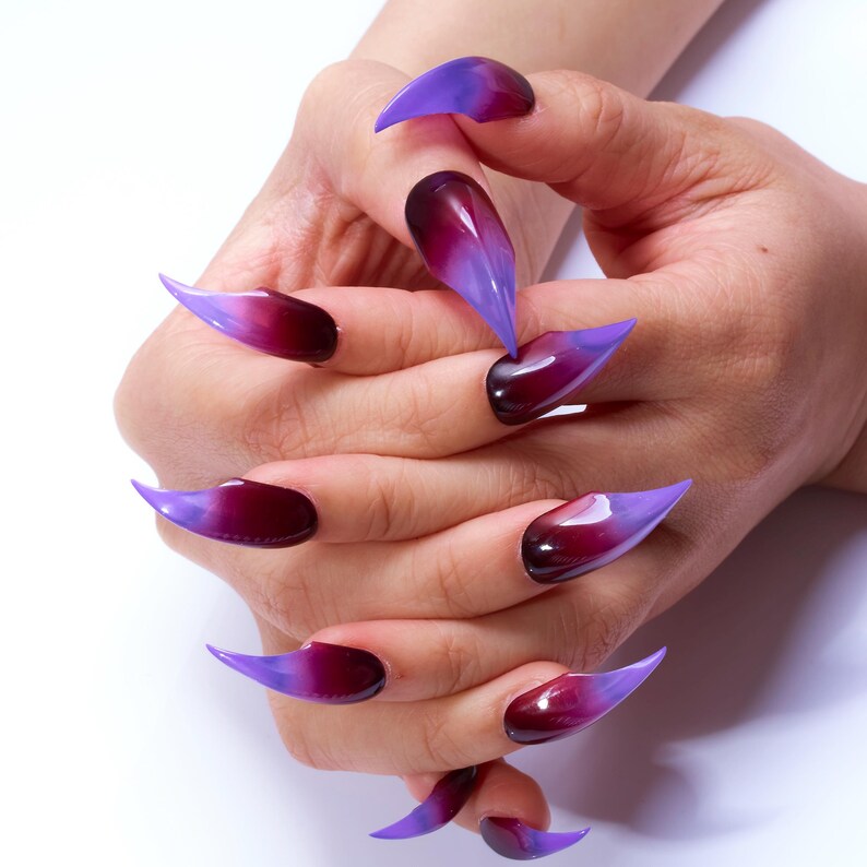 May include: Close-up of hands with long, pointed nails painted in a gradient of purple and dark red.