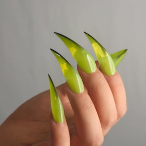 Könnte beinhalten: Eine Hand, die lange, spitze, stilettoförmige Kunstnägel zeigt. Die Nägel haben einen Farbverlauf von Limettengrün zu einem dunkleren Grün an den Spitzen, mit einem Hauch von Gelb. Der Hintergrund ist einfarbiges, neutrales Grau.