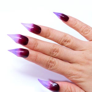 May include: A hand model wearing long, pointed, ombre nails. The nails are a light purple at the tip and fade to a dark burgundy at the base.