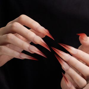 May include: Close-up of long, pointed acrylic nails with a gradient design. The nails transition from a clear base to a vibrant red at the tips. The nails are long and sharply pointed, creating a dramatic effect.