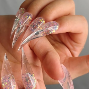 May include: Clear, pointed acrylic nails with iridescent glitter accents. The nails are long and sharp, with a clear base and glitter concentrated near the tips. The glitter reflects multiple colors, creating a sparkling effect.