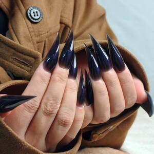 May include: A set of long, pointed, dark purple acrylic nails. The nails have a glossy finish and a subtle color gradient. The nails are on a hand, with a brown coat in the background.