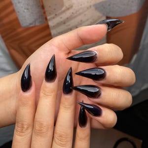 Può includere: Una mano che mostra unghie acriliche lunghe, appuntite e lucide di colore nero. Le unghie hanno una forma a stiletto affilata e il colore scuro crea un look drammatico ed elegante. Le unghie sono ben curate e riflettono la luce.