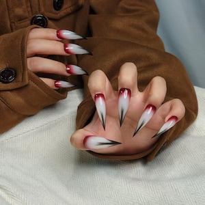 Könnte beinhalten: Lange, spitze Kunstnägel mit Farbverlauf. Die Nägel gehen von einem tiefen Rot an der Basis zu Weiß über, mit einer schwarzen Spitze. Die Nägel sind an einer Hand, mit einer braunen Jacke im Hintergrund.