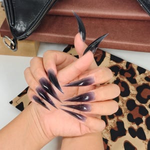 May include: Long, pointed, black and purple ombre-style artificial nails. The nails have a glossy finish and a dramatic, claw-like shape. The nails are displayed against a leopard print background.