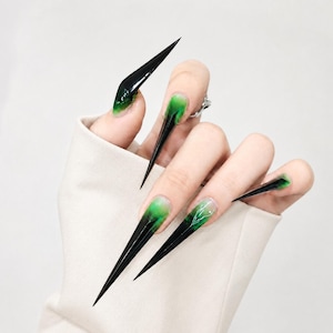 May include: Long, sharp stiletto-shaped artificial nails featuring a gradient design that transitions from a clear base to a vibrant green mid-section and ends in a solid black pointed tip. One nail displays a delicate white line art detail.