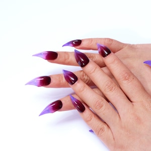 May include: A hand with long, pointed, purple and burgundy ombre nails. The nails are long and sharp, with a pointed tip.
