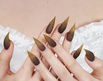 May include: A set of long, stiletto-shaped false nails with a gradient design. The nails transition from a dark brown base to a golden-yellow tip. The nails are displayed on a person's hands.