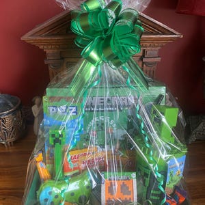 May include: A green gift basket wrapped in cellophane and tied with a large green bow. The basket contains various Minecraft-themed items, including sweets, toys, and a Creeper-shaped item. The basket sits on a wooden surface.