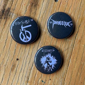 May include: Three circular pin-back buttons with black backgrounds and white graphics. One button features a hammer and peace symbol with the text "PHYSIQUE". Another displays the text "PHYSIQUE" in a stylized font. The third shows a figure.