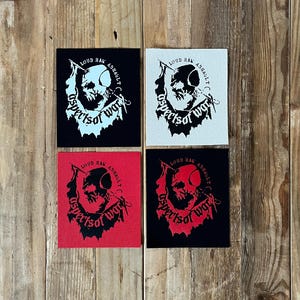 Peut inclure: Quatre écussons carrés avec un graphisme de crâne et le texte "LOUD RAK ASSAULT aspects of war". Les écussons sont noirs, blancs et rouges, avec le crâne et le texte en couleurs contrastées. Les écussons sont présentés sur une surface en bois.