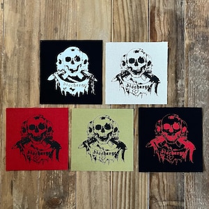 Pode incluir: Cinco patches quadrados com um desenho de caveira e a palavra "Discharge". Os patches são em preto, branco, vermelho e verde-oliva. O desenho da caveira é em preto ou branco, dependendo da cor do patch.