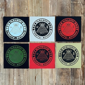 May include: Six square fabric patches featuring a circular design with a pyramid, an eye, and the text PARANOID and PNDFTW. The patches come in black with white ink, white with black ink, black with red ink, black with green ink, red with black ink, and olive with black ink.