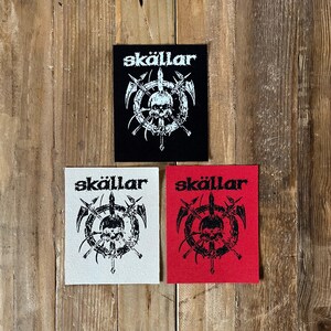 May include: Three patches with the word "skällar" above a skull and weapon design. One patch is black, one is white, and one is red. The patches are square and have a stitched border. The background is a wooden surface.