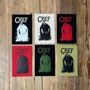 May include: Six rectangular patches with the word "GRIEF" above a stylized figure. The patches are black, off-white, red, and olive green, each with a different color figure. The patches are arranged on a wooden surface.