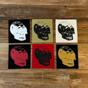 Op de afbeelding: Zes vierkante kunstdrukken met schedelontwerpen. Elke schedel heeft een andere kleur, waaronder wit, zwart, rood en goud, tegen verschillende gekleurde achtergronden. De prints zijn gerangschikt op een houten oppervlak.