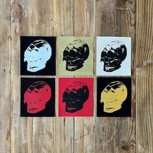 Könnte beinhalten: Sechs quadratische Aufnäher mit Totenkopfmotiven. Jeder Aufnäher zeigt einen Totenkopf in einer anderen Farbe: Weiß, Schwarz, Rot und Gelb, vor schwarzen, roten, olivfarbenen und weißen Hintergründen. Die Aufnäher sind auf einer Holzoberfläche angeordnet.