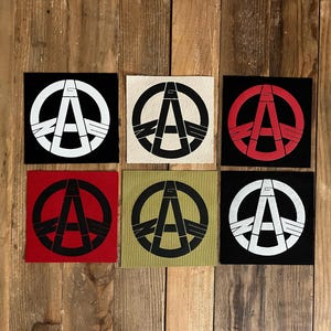 May include: Six square patches with a peace symbol design. The patches feature a black, white, red, and olive green color scheme. The peace symbol is combined with the letter "A" in a bold, graphic style. The patches are arranged on a wooden surface.