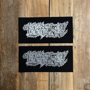 May include: Two black rectangular patches with white ornate text. The text appears to be a stylized word or phrase. The patches are on a wooden surface.
