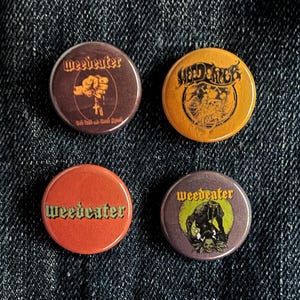 May include: Four round pinback buttons with the band name "Weedeater" and other graphics. The buttons are in shades of brown, orange, and purple. One button has a fist graphic, another has a skull graphic.