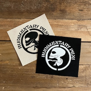 May include: Two square patches featuring a skull design and the text "RUDIMENTARY PEN". One patch is black with a white design, the other is beige with a black design. The patches are displayed on a wooden surface.