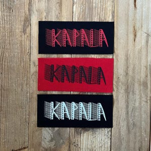 May include: Three rectangular patches with the word "KAPALA" printed on them. The top patch is black with red text, the middle patch is red with black text, and the bottom patch is black with white text. The patches are on a wooden surface.