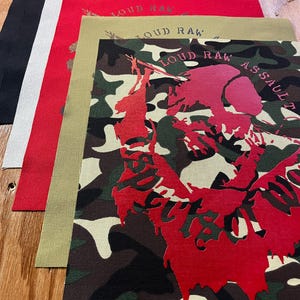 May include: A stack of five rectangular fabric patches featuring the text LOUD RAW. The top patch has a camouflage pattern with a large red graphic, while the others are solid black, white, red, and olive green with the same text printed on them.