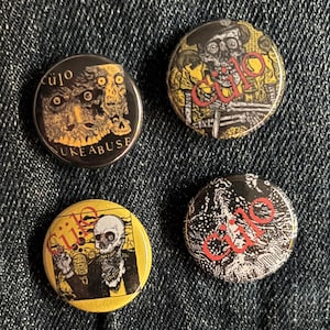 May include: Four round buttons with skull graphics and the word "cujo" in red. One button has the text "NUKE ABUSE". The buttons are black, yellow, and white, and are set against a blue denim background.