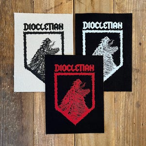 Könnte beinhalten: Drei Stoffaufnäher mit dem Wort "DIOCLETIAN" in fetter Schrift. Jeder Aufnäher zeigt eine detaillierte Illustration eines brüllenden Wesens. Ein Aufnäher ist weiß mit schwarzen Grafiken, ein anderer ist schwarz mit weißen Grafiken und der dritte ist schwarz mit roten Grafiken.