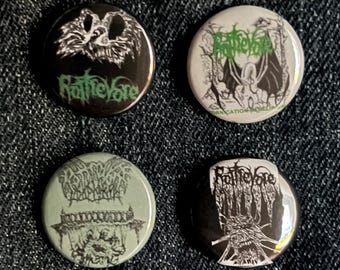 May include: Four round metal pinback buttons with black and white graphics and the word "Rattlevore" in green. The buttons feature various illustrations of creatures and text on a dark denim background.
