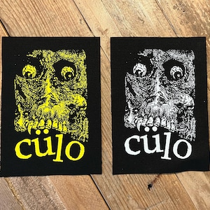May include: Two black rectangular patches with a skull design. One patch features a yellow skull with the word "cülo" in yellow. The other patch has a white skull and the word "cülo" in white.