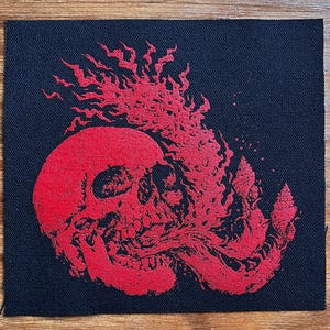 May include: A square black fabric patch featuring a screen-printed graphic in bright red ink. The design depicts a human skull with flames rising from the top and a long, flowing tongue-like shape extending from the mouth area toward the right side.
