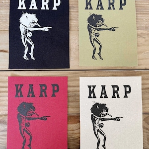 Può includere: Quattro toppe rettangolari con la scritta "KARP" in lettere bianche o nere. Ogni toppa presenta un'illustrazione stilizzata in bianco e nero di una creatura che indica. Le toppe sono in nero, verde oliva, rosso e bianco sporco.