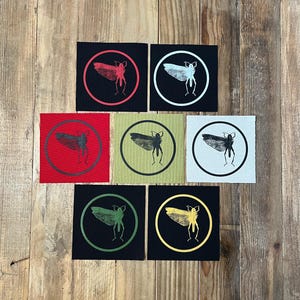 Puede incluir: Un conjunto de siete parches cuadrados con ilustraciones de insectos. Cada parche presenta un diseño circular con un gráfico detallado de insectos en varios colores, incluyendo rojo, blanco, negro, verde y amarillo. Los parches están sobre un fondo de madera.