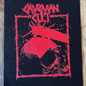 May include: Black patch with red graphic art. The design features the text "CAVEMAN CULT" in a jagged font above a red illustration of a skull and insect. The patch has a black background and a red border.