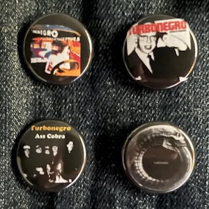 Puede incluir: Cuatro botones redondos negros con varios diseños. Un botón presenta el texto "Turbonegro" en rojo y blanco, con una imagen en blanco y negro. Otro botón tiene el texto "Turbonegro Ass Cobra". Los otros dos botones tienen imágenes.