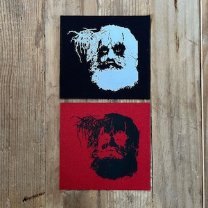 May include: Two square patches with a stylized portrait. One patch is black with a light blue image, the other is red with a black image. The image is a bearded face with a crown of thorns.