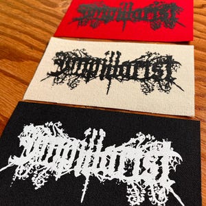 May include: Four rectangular fabric patches featuring the text "Singillarist" in a stylized, ornate black metal font. The patches come in four distinct color combinations: black text on olive, black text on red, black text on cream, and white text on black.