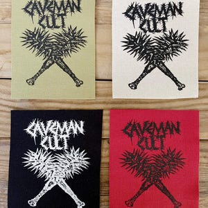 Puede incluir: Cuatro parches de tela con el texto "CAVEMAN CULT" en una fuente puntiaguda. Cada parche presenta un gráfico en blanco y negro de un objeto puntiagudo y huesos cruzados. Los parches son de color verde oliva, blanco roto, negro y rojo.