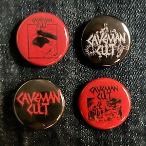 May include: Four round buttons with the text "CAVEMAN CULT" in a gothic font. Two buttons are red with black graphics, and two are black with white graphics. The buttons have various illustrations.