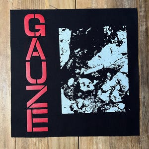 May include: Black square patch with the word "GAUNE" in red block letters on the left side. A light blue and black graphic of a skull and abstract shapes is on the right. The patch is on a wooden surface.