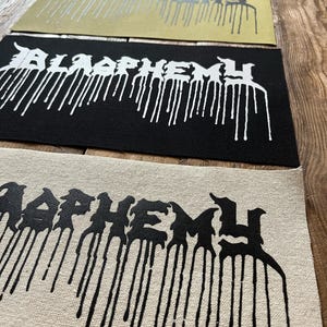 Peut inclure: Plusieurs écussons rectangulaires avec le mot "BLASPHEMY" en police gothique, avec un effet de peinture dégoulinante. Les écussons sont de différentes couleurs, dont le noir, le beige et l'or, avec du texte blanc ou argenté.
