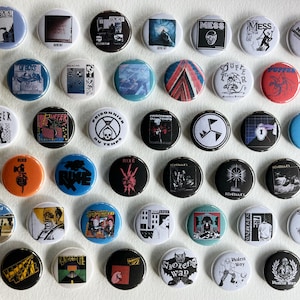 以下が含まれることがあります： 様々なデザインとテキストが施された、36個の丸いピンバッジのコレクション。ピンバッジは、黒、白、青、赤、オレンジなど、さまざまな色を使用しています。イラストが描かれたものもあれば、「MESS」や「RIXE」などのテキストが表示されているものもあります。