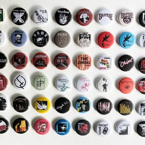 以下が含まれることがあります： 様々なデザインとテキストが施された丸いピンバッジのコレクション。バッジは、黒、白、赤、黄色など、さまざまな色を使用しています。一部のバッジには、バンド名、イラスト、グラフィックデザインが表示されています。バッジは白い表面に配置されています。
