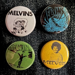 Può includere: Quattro spille rotonde con il nome della band "Melvins". Le spille presentano varie grafiche, tra cui loghi della band, illustrazioni e un ritratto. Le spille sono in bianco, nero, verde e giallo.