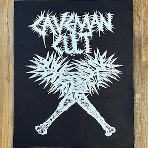 Puede incluir: Parche negro con texto y gráfico blancos. El texto dice "CAVEMAN CULT" en una fuente puntiaguda sobre una imagen estilizada de dos garrotes cruzados con cabezas puntiagudas y mangos de hueso. El parche es rectangular.